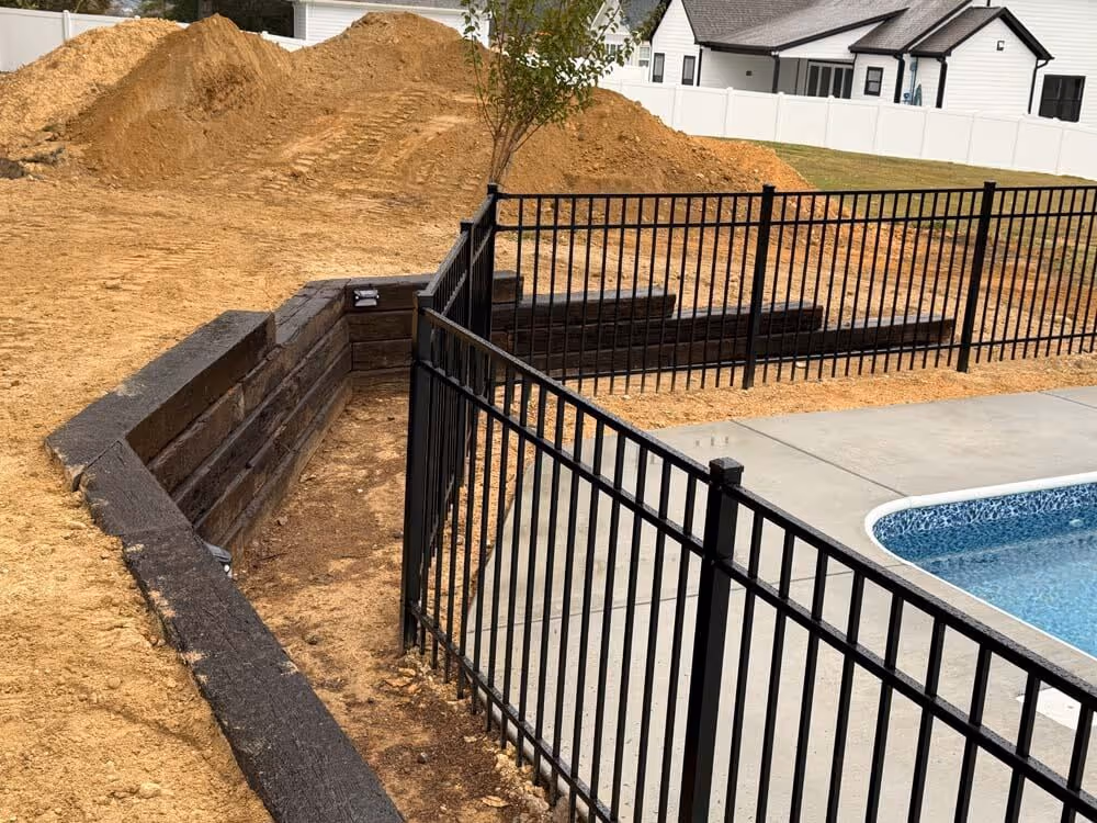 A backyard scene with a black metal fence surrounding a swimming pool and a retaining wall made of dark wood with piles of dirt behind it.