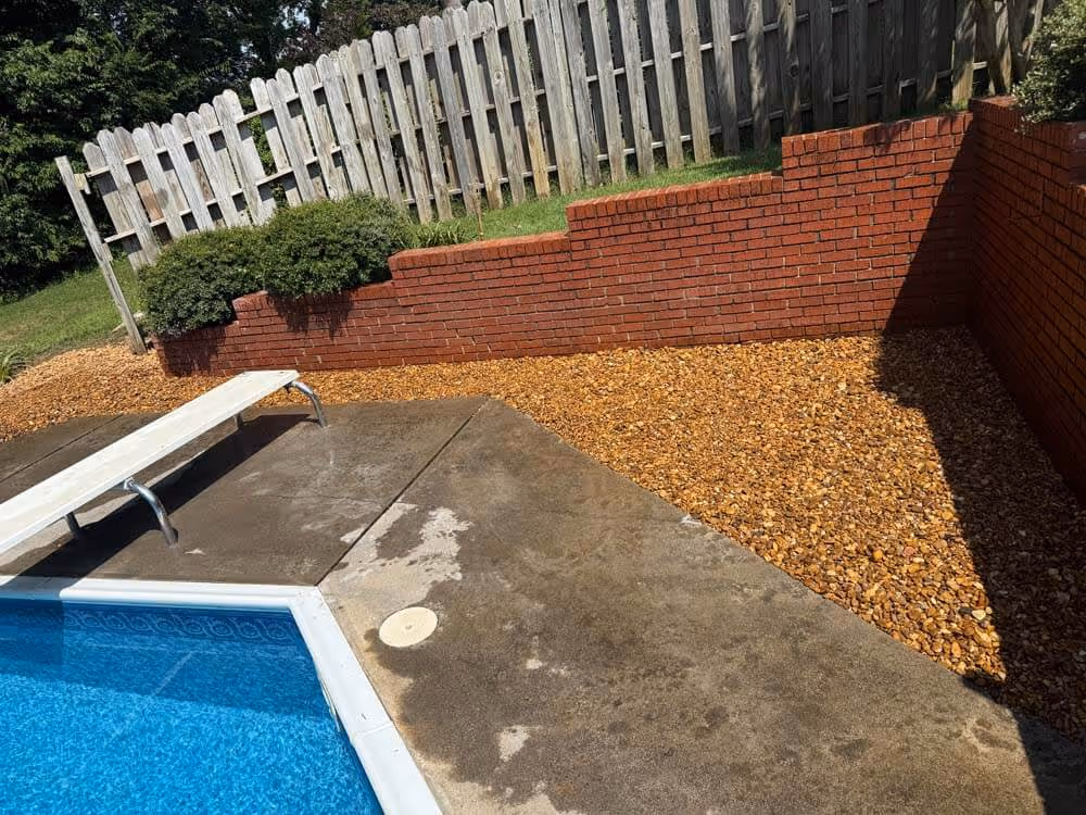 Backyard swimming pool area with a white diving board, concrete pool deck, red brick retaining wall, and gravel landscaping against a wooden fence.