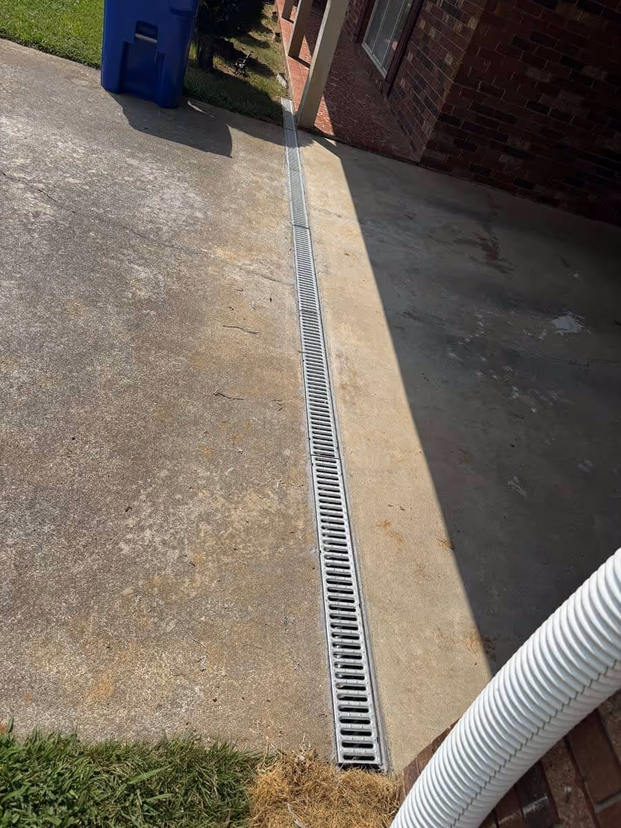 Concrete driveway with a long metal drainage grate running down the center beside a brick house and a white downspout.