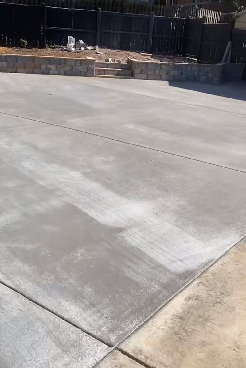 Concrete driveway with visible cleaning or resurfacing marks, bordered by dirt and a stone retaining wall with steps and a wooden fence in the background.