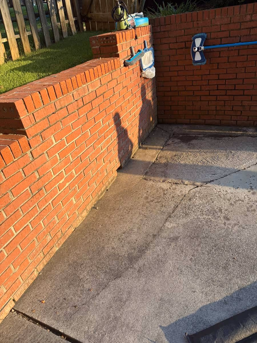 Corner of a brick wall next to a concrete patio with two blue cleaning brushes hanging on the wall.
