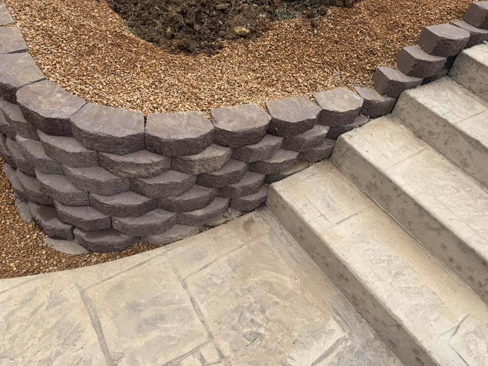 Curved stacked stone retaining wall next to concrete steps and beige gravel ground.