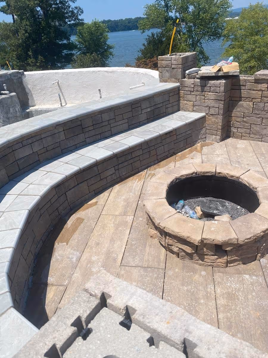 Curved stone seating area surrounding a circular stone fire pit on a patio with a lake and trees in the background.