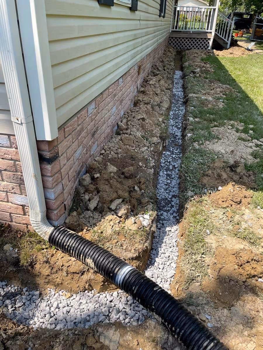 House foundation trench filled with gravel and a black corrugated drainage pipe connecting to a downspout.