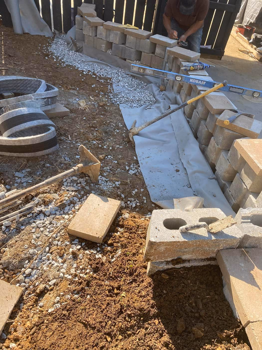 Construction site with a partially built cinder block wall, gravel spread on a fabric liner, and various masonry tools including a level, hammer, and trowel.