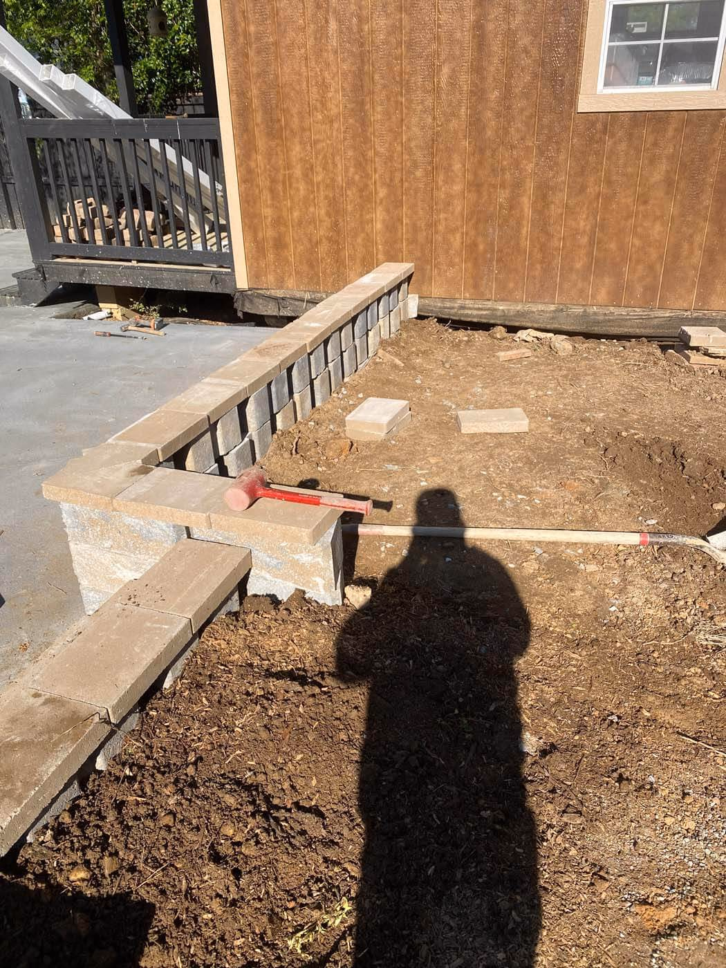 Construction site showing a partially built stone retaining wall next to bare soil and a wooden building.