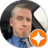 Man with light skin and short hair wearing a blue shirt and tie inside a car with an orange Google review badge.