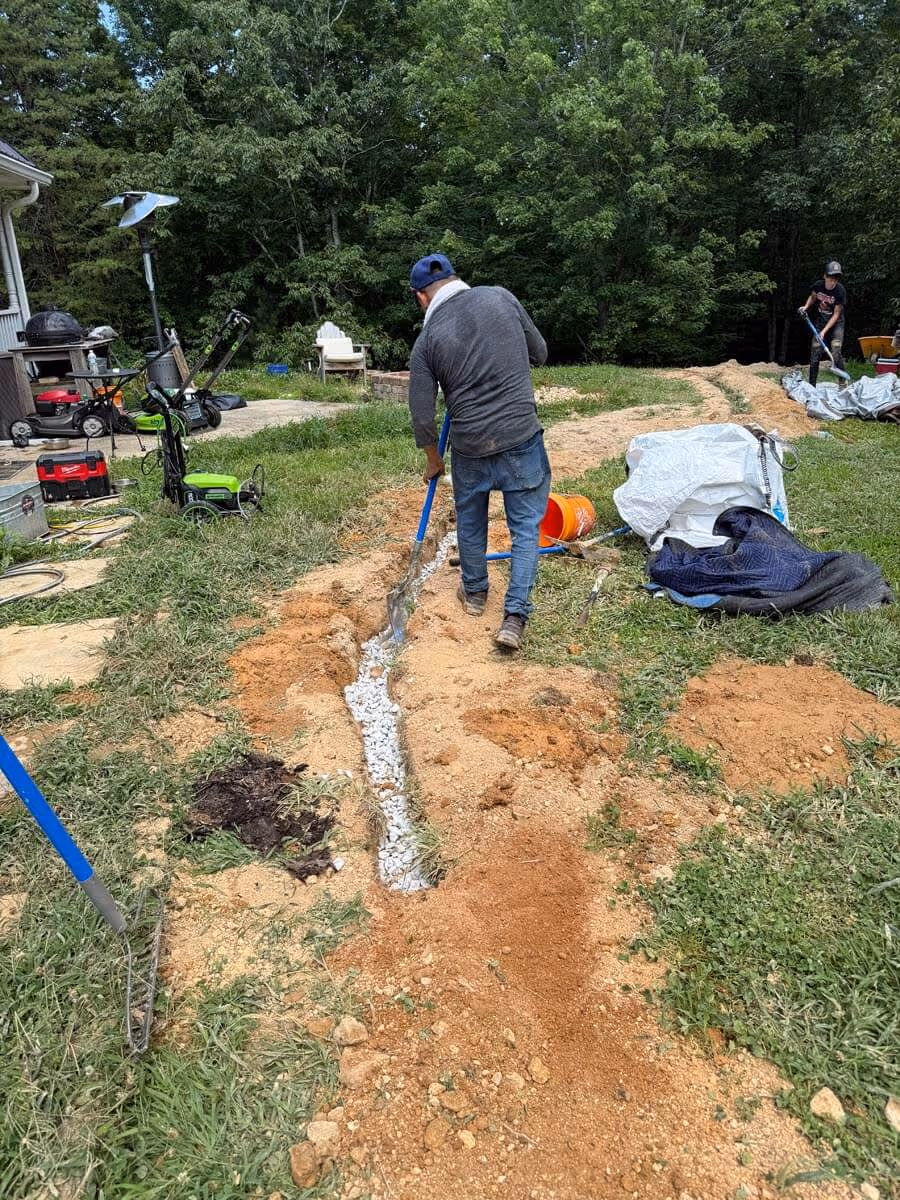 Man in work clothes filling a trench with gravel using a shovel in a grassy yard with trees and various tools nearby.