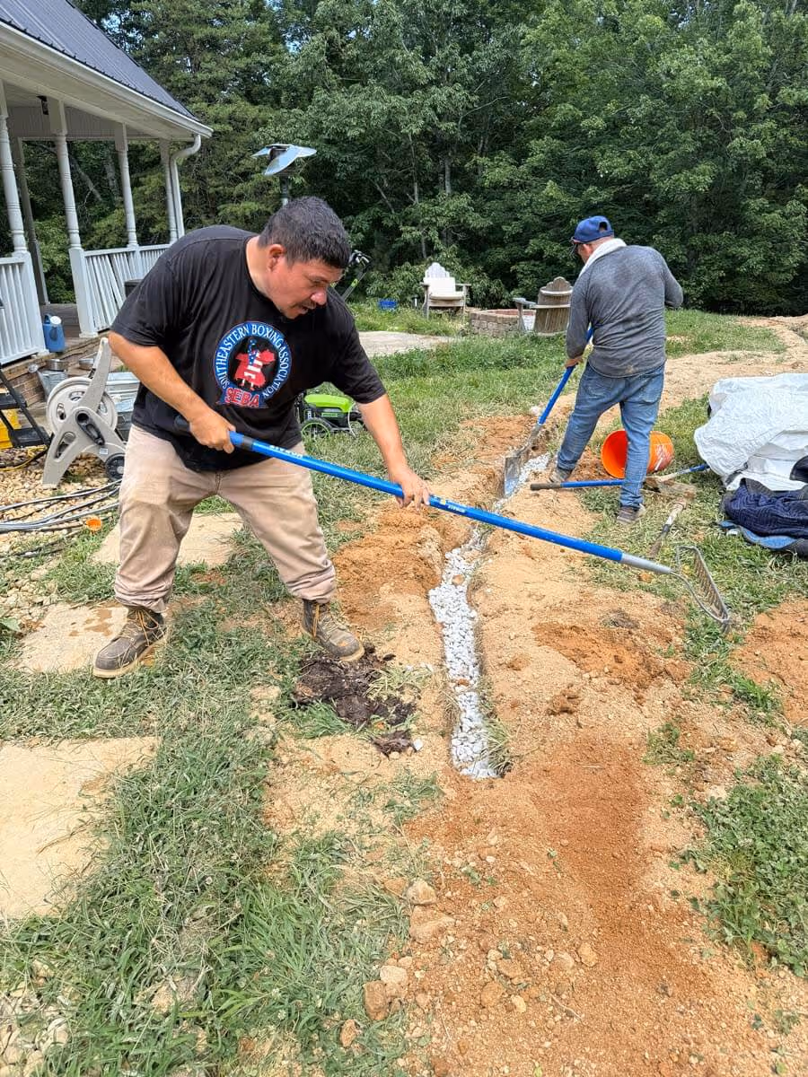 Two men working outdoors with rakes, spreading gravel and dirt in a trench near a house with trees in the background.
