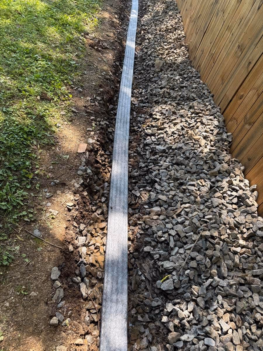 A narrow trench with a drainage strip laid in the middle, bordered by grass on one side and a wooden fence with gravel on the other.
