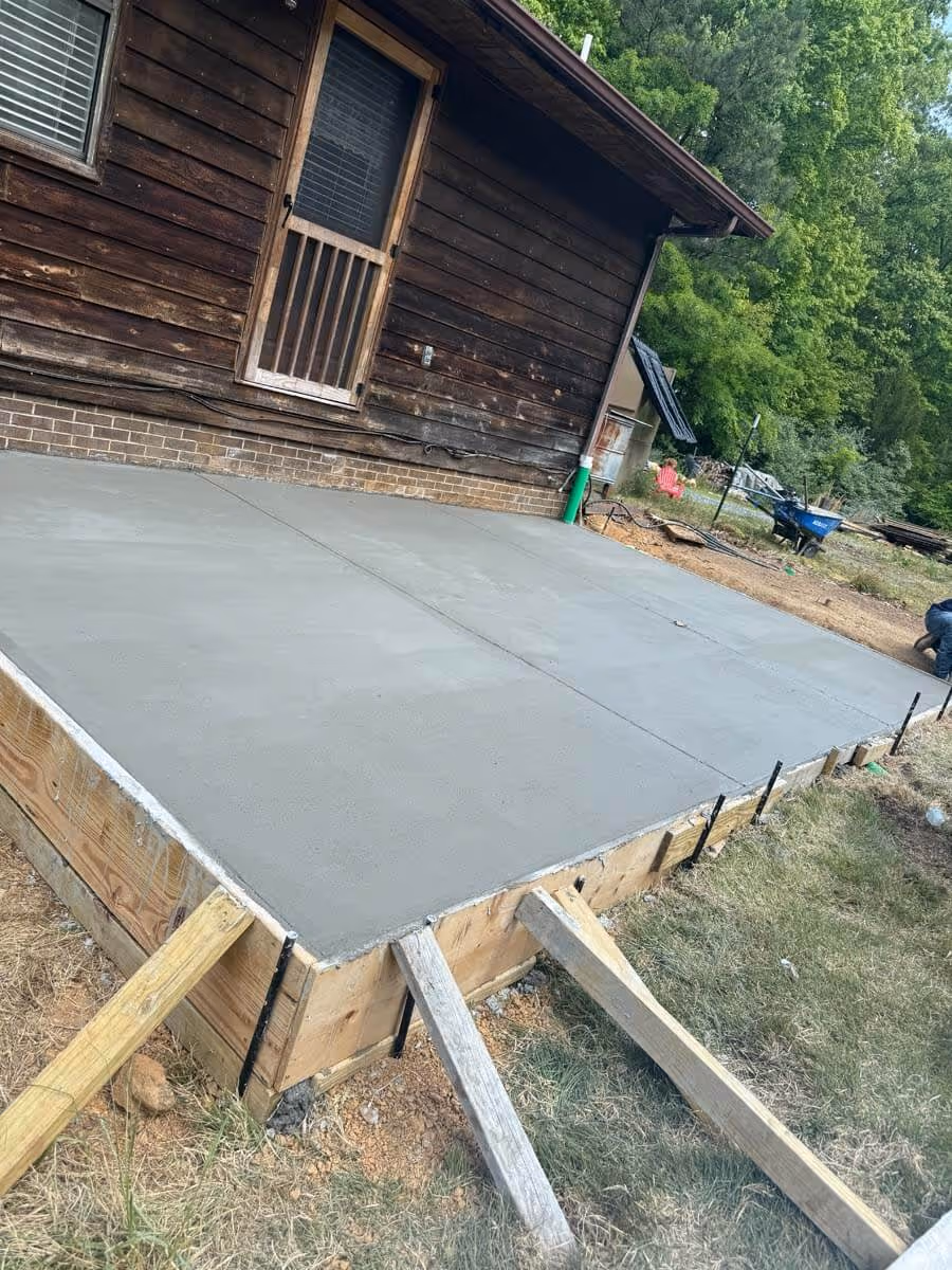 Newly poured smooth concrete slab next to a wooden house with surrounding greenery and construction tools nearby.