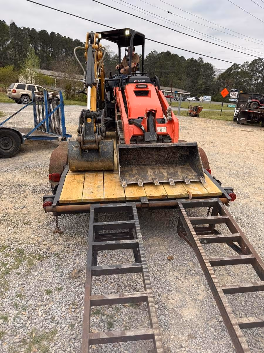 Orange Kubota mini excavator loaded on a trailer with metal ramps on a gravel surface during overcast weather.