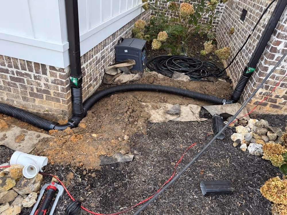 Outdoor corner of a brick house with black corrugated drainage pipes installed at the base and garden soil partially dug up.