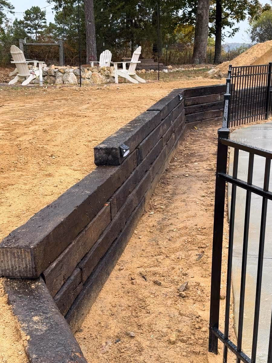 Outdoor seating area with four white Adirondack chairs around a stone firepit, next to a wooden retaining wall and black metal fence on sandy ground.