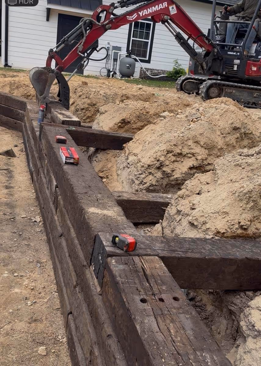 Red Yanmar excavator digging near a wooden retaining wall under construction with piles of dirt beside it.
