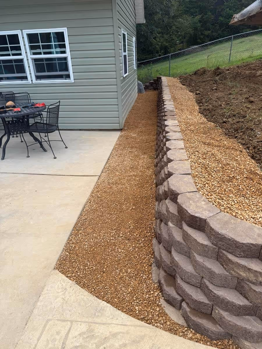 Side of a house with green siding, a small metal patio table with chairs on a concrete patio, and a gravel pathway bordered by a tiered stone retaining wall and soil bed.