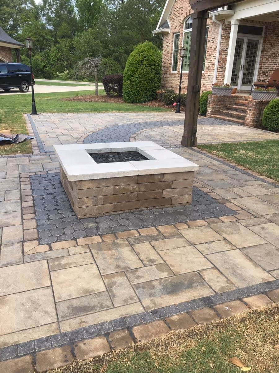 Square stone fire pit surrounded by a multi-patterned paved patio in a backyard with brick house and greenery.