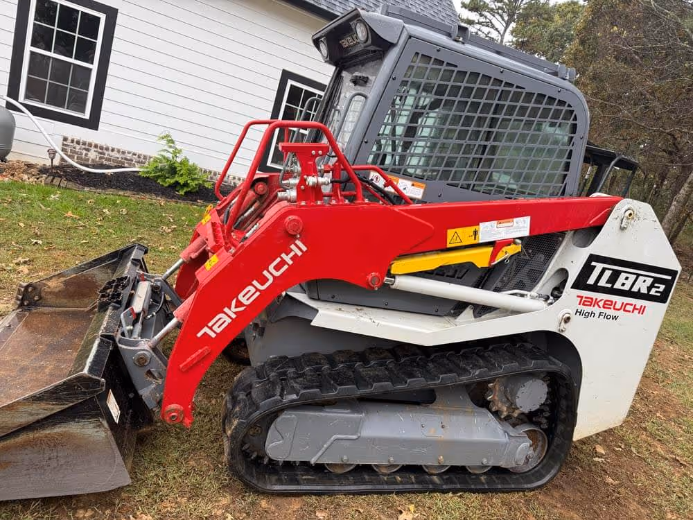 Takeuchi TL8R2 compact track loader in red, white, and gray parked on grass near a white house.