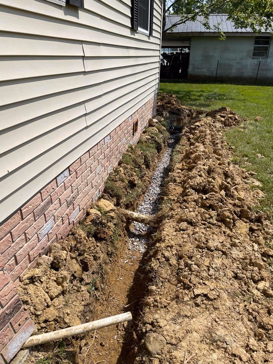 Trench dug alongside a house foundation with gravel at the bottom and PVC pipes partially installed.