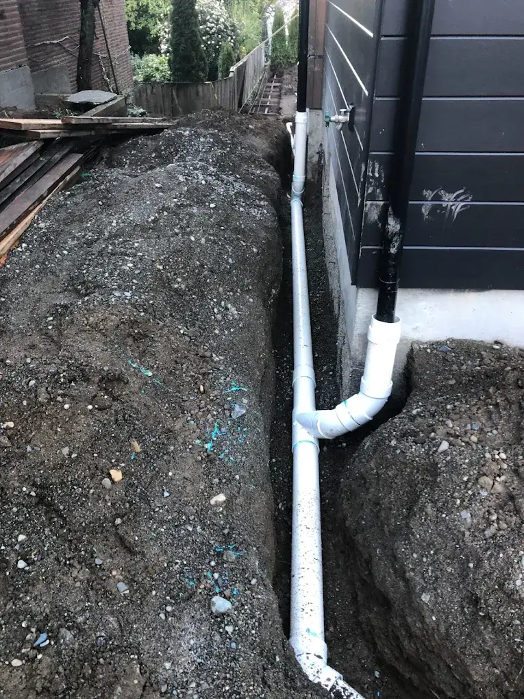 White PVC drainage pipes installed in a narrow trench beside a dark exterior building wall with soil piled on the other side.