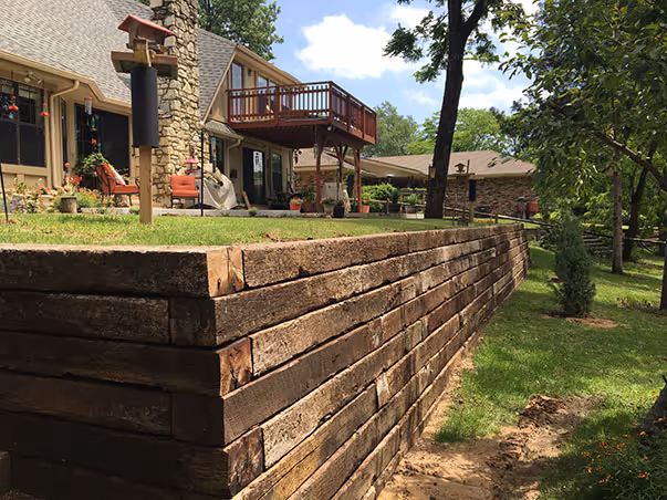Wooden retaining wall supporting a garden area next to a house with a stone chimney and a raised wooden deck.